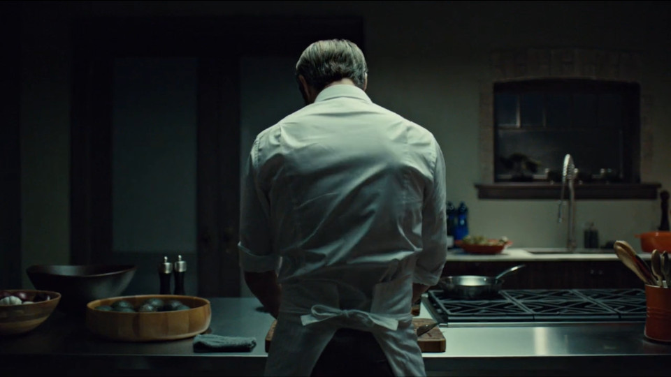 In a dark kitchen, Hannibal's white shirt and apron is illuminated from above. There are pieces of cookward around him, and he is leaning over a cutting board.